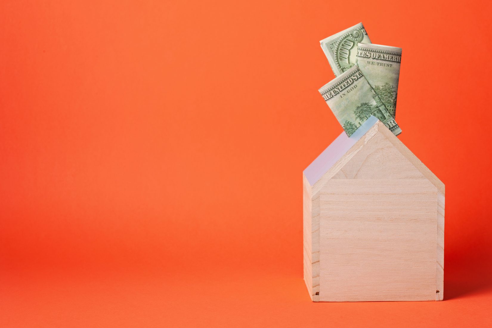 Wooden toy house with money sticking out of the top, set against an orange background